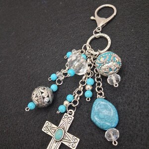 May include: A silver-toned keychain featuring a lobster clasp and a split ring. The charm assembly includes a silver cross with a central turquoise inlay, a large turquoise-colored stone, a filigree bead, and several clear and blue beads.