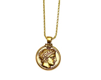 May include: A gold chain necklace with a round pendant featuring a profile of a person with a laurel wreath. The pendant is a replica of an ancient Greek coin.