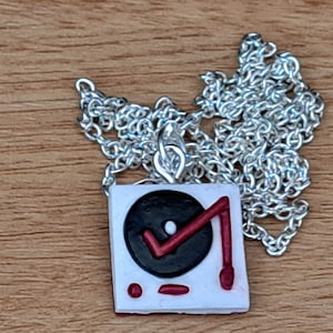 May include: A silver chain necklace with a white pendant shaped like a record player. The record is black with a red needle and a white dot in the centre.