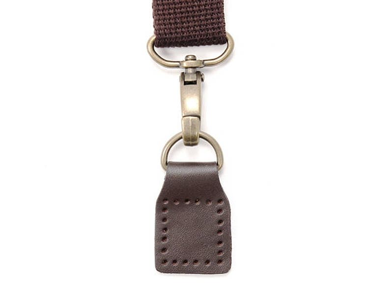 May include: A brown leather strap with a brass hook and loop closure. The strap is attached to a brown leather loop with a stitched edge.