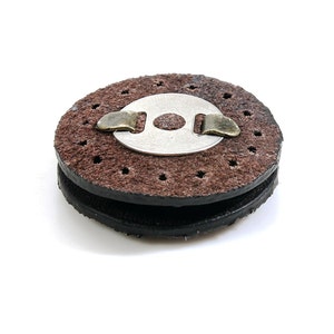 May include: A brown leather disc with a metal ring and a small metal clip. The disc has a series of small holes around the edge.