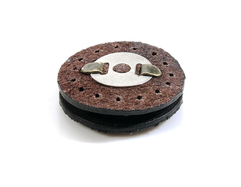 May include: A brown leather disc with a metal ring and a small metal clip. The disc has a series of small holes around the edge.