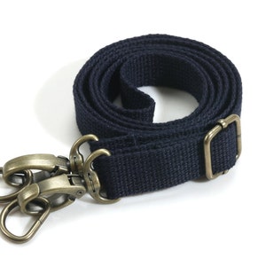 May include: A navy blue canvas strap with antique brass-colored metal hardware. The strap is rolled up, showing its length and texture. The hardware includes two swivel clasps and an adjustable buckle, suitable for bags or other accessories.