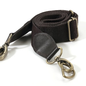 May include: A brown canvas camera strap with two gold metal clasps. The strap is rolled up and the clasps are visible.