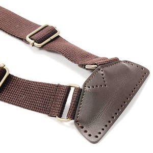 May include: Brown canvas strap with a leather patch and two metal buckles. The strap is designed for a bag or other accessory.