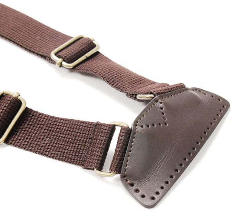 May include: Brown canvas strap with a leather patch and two metal buckles. The strap is designed for a bag or other accessory.