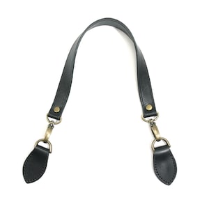 May include: A black leather bag strap featuring a curved handle design with brass-toned metal hardware. The strap includes two swivel lobster clasps attached to two separate black leather tabs, each secured with a single brass-toned rivet.