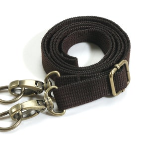 May include: A dark brown canvas strap with antique brass-colored metal hardware. The strap is coiled, showing a buckle and two swivel snap hooks. The hardware is attached to the strap, ready for use.