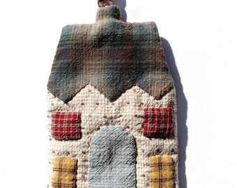 May include: A small, house-shaped key chain made of patchwork fabric. The house has a brown and green plaid roof, a white body with red and yellow plaid windows, and a gray door. A silver key ring is attached to the bottom.