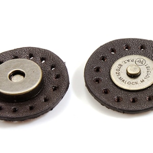 Puede incluir: Dos cierres magnéticos de cuero marrón con detalles metálicos. El cierre de la derecha tiene el texto "MALOCK. M. 10131871 PAT. 8702412" en el detalle metálico.