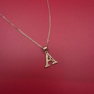 Puede incluir: Un collar dorado con un colgante con la forma de la letra "A". El colgante presenta un diseño calado detallado. El collar se muestra sobre un fondo rojo liso.