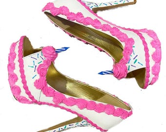 May include: A pair of white high-heeled shoes decorated to look like a birthday cake with pink frosting and colorful sprinkles.