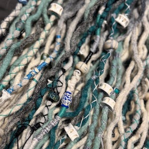 May include: Close-up of handmade dreadlocks in shades of teal, gray, and cream. The dreads are adorned with wooden beads and decorative wraps. The texture is thick and fibrous, with a bohemian aesthetic.