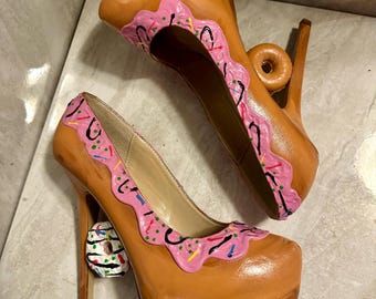 May include: A pair of high-heeled shoes with a platform sole. The shoes are painted to look like donuts, with pink frosting, sprinkles, and a chocolate glaze. The heels are also shaped like donuts.