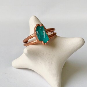 Op de afbeelding: Een koperen ring met een turquoise groene edelsteen in het midden. De ring ligt op een witte keramische ringhouder.