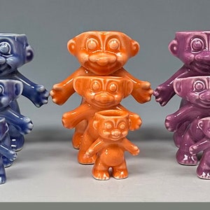May include: A set of nine ceramic figurines stacked in groups of three. The figurines are all in different colors, including pink, green, blue, orange, purple, and yellow. Each figurine has a smiling face and a simple, rounded body.