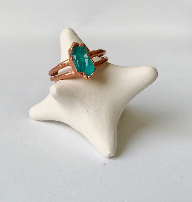 Op de afbeelding: Een koperen ring met een turquoise groene edelsteen in het midden. De ring ligt op een witte keramische ringhouder.