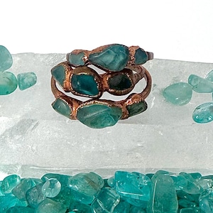 May include: A copper ring featuring a series of raw, teal-colored stones embedded along the band. The metal has a textured, metallic finish, and the stones are irregular in shape, displaying varying shades of blue-green throughout the design.
