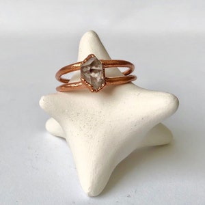 Op de afbeelding: Een ring van ros&eacute;goud met een helder kwartskristal in het midden. De ring is dubbel gevat en zit op een witte stervormige ringhouder.