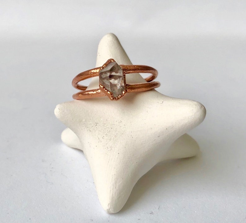 Op de afbeelding: Een ring van ros&eacute;goud met een helder kwartskristal in het midden. De ring is dubbel gevat en zit op een witte stervormige ringhouder.