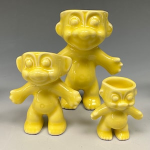 May include: Set of three yellow ceramic egg cups shaped like cartoon figures. The egg cups are different sizes and have a whimsical, retro style.