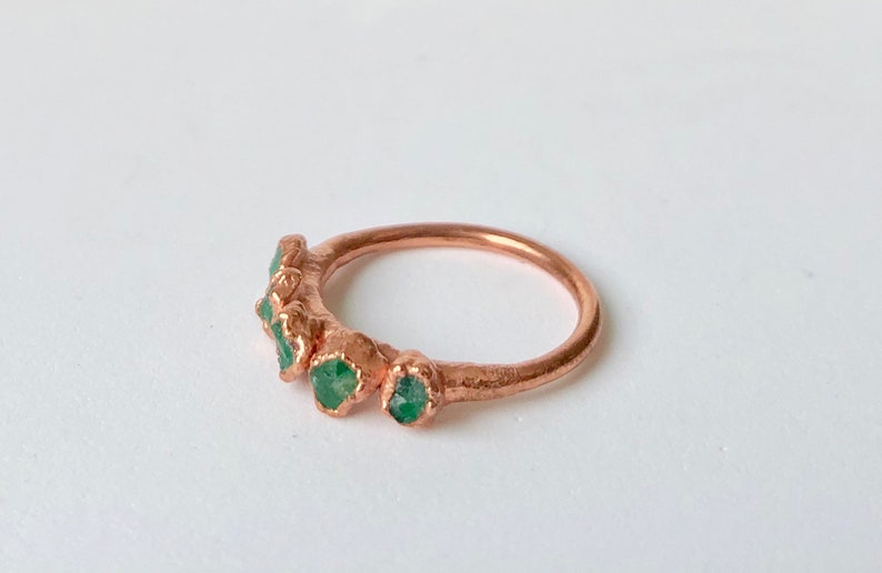 Op de afbeelding: Een koperen ring met vijf groene edelstenen die in de band zijn gezet. De ring is gemaakt van gehamerd koper en heeft een rustieke uitstraling.