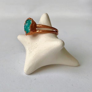 Op de afbeelding: Een koperen ring met een turquoise edelsteen. De ring ligt op een witte keramische ringhouder.