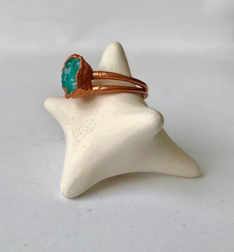 Op de afbeelding: Een koperen ring met een turquoise edelsteen. De ring ligt op een witte keramische ringhouder.