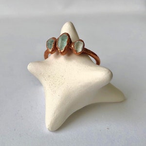 Op de afbeelding: Een koperen ring met drie lichtblauwe edelstenen die in de band zijn gezet. De ring wordt getoond op een witte stervormige ringhouder.