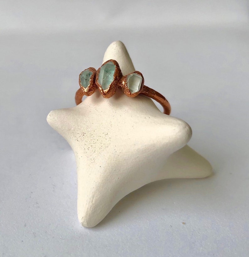 Op de afbeelding: Een koperen ring met drie lichtblauwe edelstenen die in de band zijn gezet. De ring wordt getoond op een witte stervormige ringhouder.