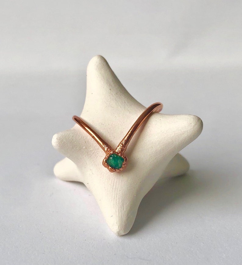 Op de afbeelding: Een koperen ring met een groene edelsteen in het midden. De ring is V-vormig en staat op een witte keramische standaard.