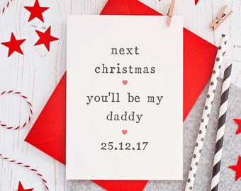 Puede incluir: Una tarjeta blanca con el texto "next christmas you'll be my daddy 25.12.17" con un símbolo de corazón rojo entre "christmas" y "you'll" y entre "daddy" y la fecha. La tarjeta está sobre un sobre rojo y una superficie gris. Hay estrellas rojas y una cuerda roja y blanca esparcidas alrededor de la tarjeta.