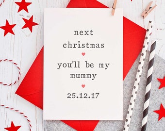 Puede incluir: Una tarjeta blanca con corazones rojos y el texto "next christmas you'll be my mummy 25.12.17" sobre un fondo de madera blanco con cuerda roja y blanca, estrellas rojas y pajitas rayadas grises y negras.