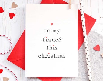 May include: A white card with the text "to my fiancé this christmas" in black font with small red hearts scattered throughout the text. The card is on top of a red envelope and a gray surface.