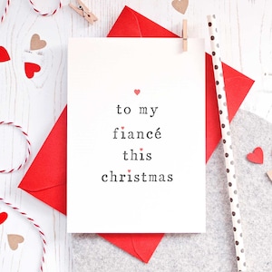 May include: A white card with the text "to my fiancé this christmas" in black font with small red hearts scattered throughout the text. The card is on top of a red envelope and a gray surface.