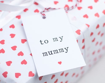 Puede incluir: Una caja de regalo blanca con un patrón de corazones rojos y una etiqueta de regalo blanca que dice "a mi mami" con un pequeño corazón rojo en la parte inferior.