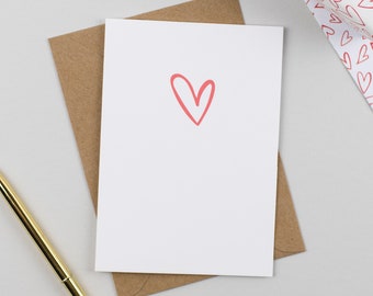 Puede incluir: Una tarjeta de felicitación blanca con un corazón rojo dibujado a mano, colocada sobre un sobre de papel kraft marrón. Un bolígrafo metálico dorado se encuentra en diagonal, y se ve una caja de regalo envuelta en papel blanco con corazones.