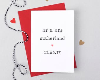 May include: A white card with red and black trim, with the text "mr & mrs sutherland 11.02.17" in black typewriter font. The card is surrounded by small red and brown hearts.