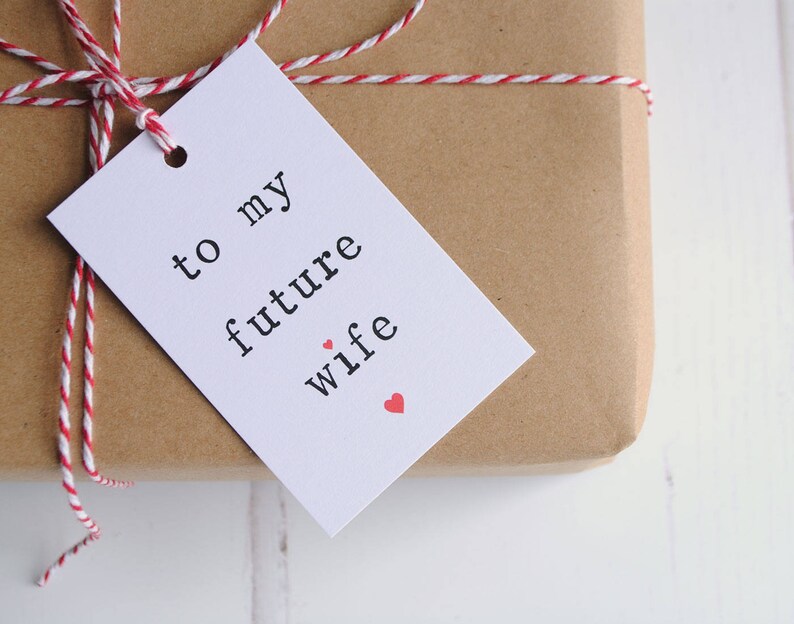 May include: A white gift tag with black text that reads "to my future wife" with two small red hearts. The tag is tied to a brown paper wrapped gift with red and white twine.
