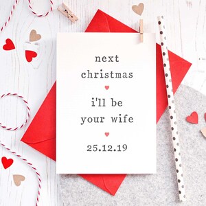 May include: White card with black text that reads "next christmas i'll be your wife 25.12.19". There are small pink hearts scattered throughout the text. The card is sitting on a red envelope.