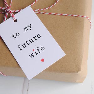 May include: White gift tag with black text that reads "to my future wife" with a small red heart. The tag is tied to a brown paper package with red and white twine.