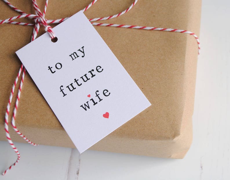 May include: White gift tag with black text that reads "to my future wife" with a small red heart. The tag is tied to a brown paper package with red and white twine.