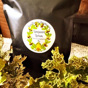 May include: A black bag with a circular label reading "Fern Flower Apothecary" and "Lungwort Lichen". The label is surrounded by a green and yellow wreath. Dried green lichen is in the foreground.