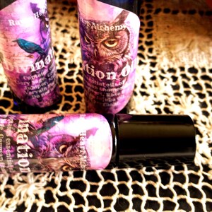 May include: Three purple essential oil bottles with black caps are displayed. The labels feature illustrations of an owl and a bird, along with the words "Divination Oil" and "Potion Oil". The bottles are on a white lace surface.
