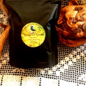 May include: A black pouch with a round label featuring a bird illustration and the text "Lungwort Lichen". The pouch is on a white lace surface, with a wooden object and a cinnamon stick visible.