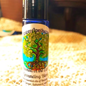 May include: A blue glass essential oil roller bottle with a black cap. The label features a tree design with the text "Flower Apotheca" and "Rooted in the Earth Grounding Blend". The blend contains essential oils of Benzoin, Ginger, Spikenard, and Sweet Almond Oil.