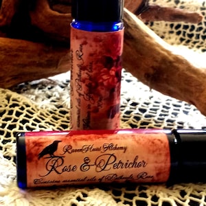 May include: Two cylindrical glass roller bottles with black caps. The labels feature a red and orange textured background with the text "Raven Heart Alchemy Rose & Petrichor" and "Contains essential oils of Patchouli, Rose".