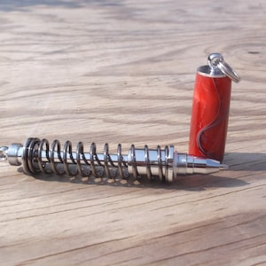 May include: A silver pen with a spring-loaded design and a red and black patterned pen cap. The pen is on a wooden surface.