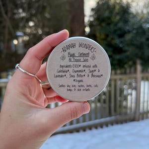 May include: A small metal tin with a white label that reads "ADAMAH WONDERS- Magic Ointment All Purpose Salve" and lists the ingredients: EVOO infused with Calendula, Chamomile, Sage & Lavender, Shea Butter & Beeswax. The label also states "*Organic" and "Soothes dry skin, rashes, burns, cuts, bumps & skin irritants".