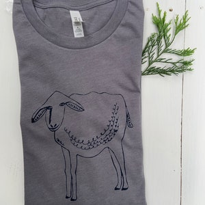 May include: A gray t-shirt with a black and white illustration of a sheep. The sheep is drawn in a simple, stylized way, with a curly tail and a friendly face.